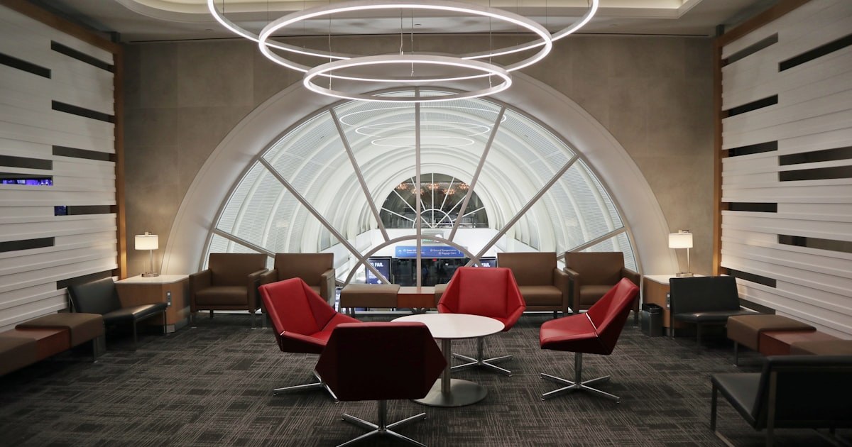 Bright modern airport lounge seating area with red chairs under a circular light fixture and arched glass window